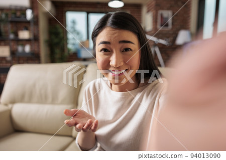 Happy young asian chineese filipino woman sitting on sofa home holding phone at screen waving hand video calling distance friend online in mobile chat app using smartphone videochat application 94013090
