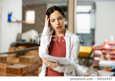 Confused female designer standing with papers in apartment during renovation 94016487