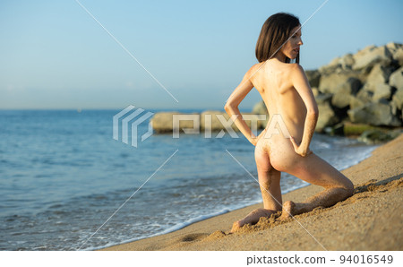 Rear view of nude young woman posing on knees on seashore 94016549