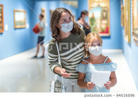 Mother and daughter in protective masks looking at paintings in halls of museum 94016896