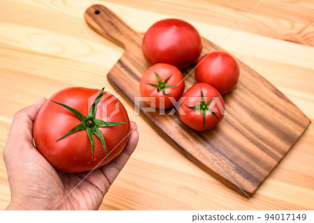 A tomato in hand and multiple tomatoes lined up on a wooden cutting board on the table 94017149