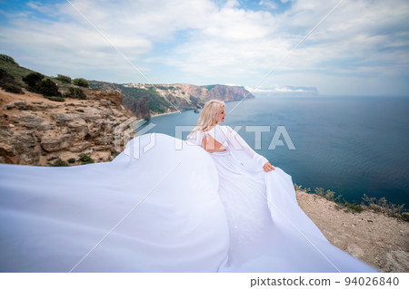 Blonde with long hair on a sunny seashore in a white flowing dress, rear view, silk fabric waving in the wind. Against the backdrop of the blue sky and mountains on the seashore. Blonde with long hair on a sunny seashore in a white flowing dress, rear view, silk fabric waving in the wind. Against the backdrop of the blue sky and mountains on the seashore. 94026840