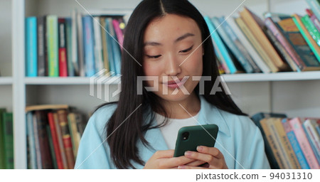 Attractive asian woman took a break between studies and using smartphone, texting message while standing in the library. University library concept. 94031310