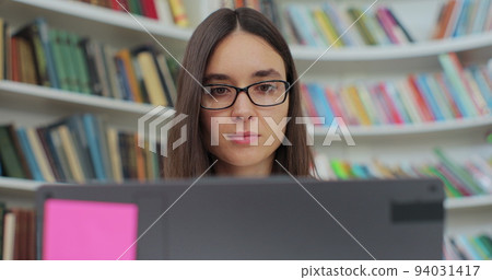 Beautiful young woman in glasses reading book and typing on computer. College student works on essay at the library. Beautiful young woman in glasses reading book and typing on computer. College student works on essay at the library. 94031417