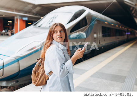 Young redhhead woman waiting train on station platform with backpack and using smart phone. Railroad transport concept, Traveler. Woman traveler walking at train station 94033033