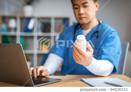 Serious adult chinese male doctor with laptop looks at jar of pills in clinic office interior. Treatment with therapist 94036531