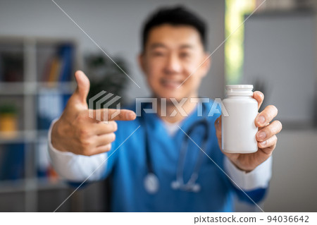 Smiling adult korean male doctor pointing finger at jar of pills in clinic office interior, selective focus Smiling adult korean male doctor pointing finger at jar of pills in clinic office interior, selective focus 94036642