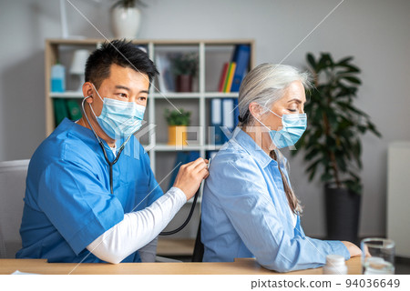 Concentrated millennial korean man doctor in protective mask with stethoscope listens to breath of elderly female 94036649