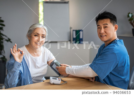 Handsome mature korean man doctor measures pressure with tonometer to senior female, patient waving hand 94036653