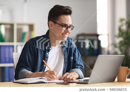 Happy millennial businessman taking notes while working with laptop in office 94036655