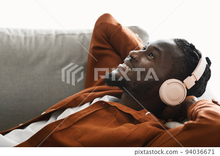 Positive black man in headphones lying on sofa and listening to music, dreaming and relaxing with hands behind head 94036671