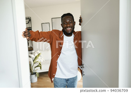 Happy african american man opening door of his new apartment and smiling at camera, standing indoors 94036709