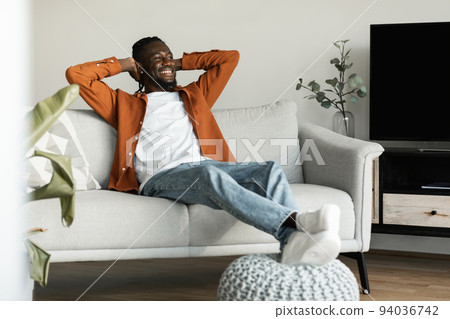 Rest concept. Happy black man sitting on comfortable couch at home in living room, leaning back with hands behind head 94036742