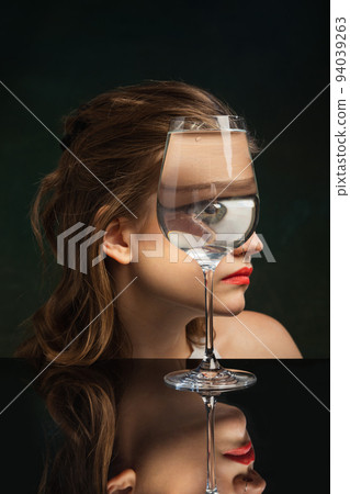 Creative art photography. Closeup girl's face through wine glass over dark background. Object distortion, optical illusion. Minimalistic contemporary art. 94039263