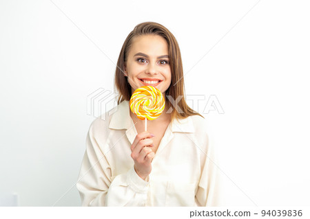 Beautiful young caucasian woman wearing a white shirt licking a lollipop on a white background Beautiful young caucasian woman wearing a white shirt licking a lollipop on a white background 94039836