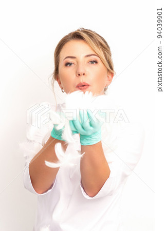 Young beautiful woman beautician in protective green gloves standing and blowing on feathers over white wall background 94039901