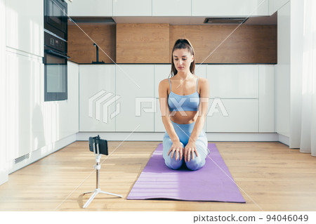 Time to train. Young beautiful fitness woman doing workout with dumbbells online with phone through video call from home, sitting on mat, quarantine, isolation 94046049