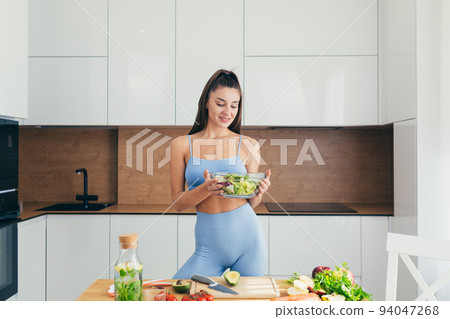 Beautiful young vegetarian woman in sports uniform prepares healthy food from vegetables at home in the kitchen Beautiful young vegetarian woman in sports uniform prepares healthy food from vegetables at home in the kitchen 94047268