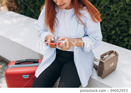 Woman with a red suitcase using smartphone in the European city. Hipster girl browsing Internet on a phone, texting and communicating outdoors. Travel concept 94052953