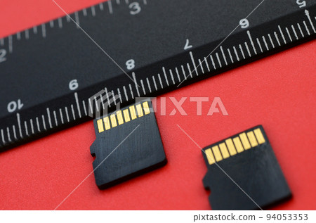Two small micro SD memory cards lie on a red background next to a black ruler. A small and compact data and information store 94053353