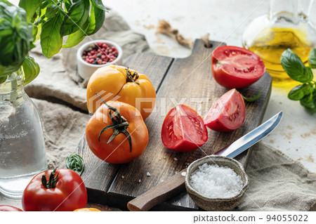 Raw tomatoes and knife on wooden cutting board surrounded by ingredients salt basil leaves and olive oil around. Rustic cooking background 94055022