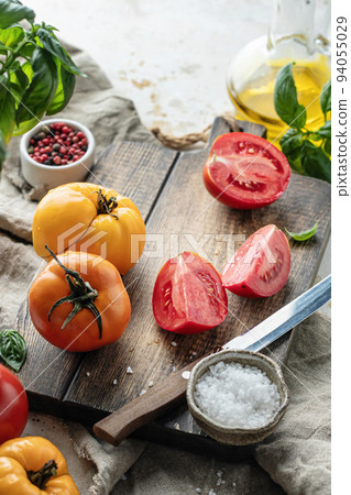 Raw tomatoes and knife on wooden cutting board surrounded by ingredients salt basil leaves and olive oil around. Rustic cooking background 94055029