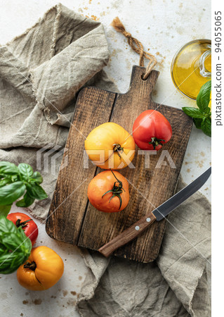 Raw tomatoes and knife on wooden cutting board surrounded by ingredients salt basil leaves and olive oil around. Top view. Rustic cooking background 94055065