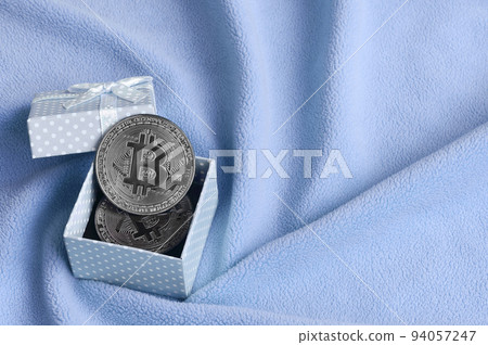 The silver bitcoin lies in a small blue gift box with a small bow on a blanket made of soft and fluffy light blue fleece fabric with a large number of relief folds The silver bitcoin lies in a small blue gift box with a small bow on a blanket made of soft and fluffy light blue fleece fabric with a large number of relief folds 94057247