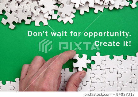 Don't wait for opportunity. Create it. The hand folds a white jigsaw puzzle and a pile of uncombed puzzle pieces lies against the background of the green surface Don't wait for opportunity. Create it. The hand folds a white jigsaw puzzle and a pile of uncombed puzzle pieces lies against the background of the green surface 94057312