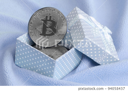 The silver bitcoin lies in a small blue gift box with a small bow on a blanket made of soft and fluffy light blue fleece fabric with a large number of relief folds The silver bitcoin lies in a small blue gift box with a small bow on a blanket made of soft and fluffy light blue fleece fabric with a large number of relief folds 94058437