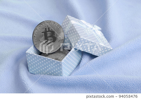 The silver bitcoin lies in a small blue gift box with a small bow on a blanket made of soft and fluffy light blue fleece fabric with a large number of relief folds The silver bitcoin lies in a small blue gift box with a small bow on a blanket made of soft and fluffy light blue fleece fabric with a large number of relief folds 94058476
