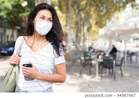 Woman in mask with smartphone walking through city streets Woman in mask with smartphone walking through city streets 94067007