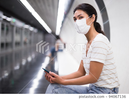 Woman in face mask with smartphone sitting on bench in subway station 94067136