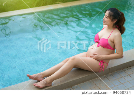 happy pregnant woman in bikini sitting on edge of swimming pool 94068724