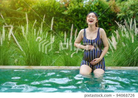 cheerful pregnant woman in swimsuit sitting on edge of swimming pool 94068726