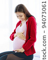Pregnant woman sits on the bed in her bedroom, caressing her belly with a happy, smiling face. 94073061