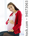 Pregnant woman sits on the bed in her bedroom, caressing her belly with a happy, smiling face. 94073063