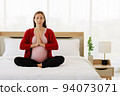Pregnant woman sits in bed in her bedroom meditating alone in peace and happiness. The new mother resting by sitting down to chill out. 94073071