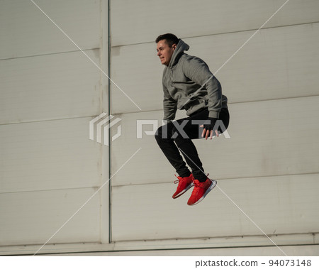 Caucasian man in hoodie jumping high with hands on knees outdoors.  94073148