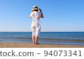 back view on a mature woman in a straw hat and white dress walks along the blue sea coast on a sunny 94073341