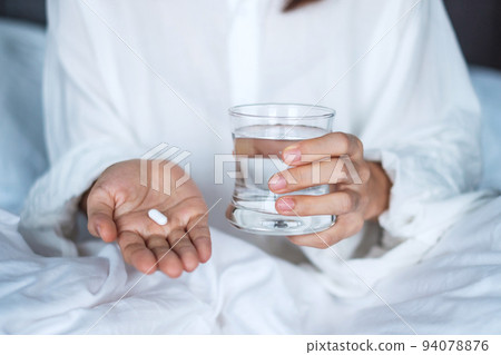 Adult woman holding pill and glass of water, female taking medicine on bed at home. Migraine, painkiller, headache, influenza, illness, sickness and healthcare concept Adult woman holding pill and glass of water, female taking medicine on bed at home. Migraine, painkiller, headache, influenza, illness, sickness and healthcare concept 94078876