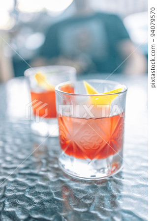 Glass of a Boulevardier cocktail with big ice cube and orange zest at bar, outdoors 94079570