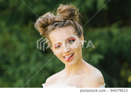 A cheerful funny young woman or girl with bright makeup and a funny hairstyle squinted one eye and shows a smiling tongue 94079623