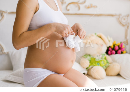 A pregnant woman holds white booties in her hands. A young woman in white underwear for pregnant women is waiting for the birth of a child. Pregnancy, Motherhood, the concept of the Mother's Day 94079636