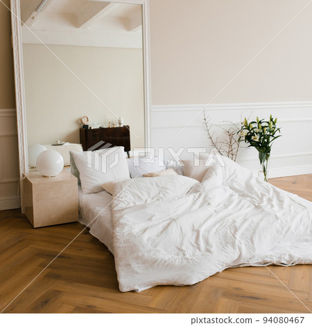 A double bed with white linens and beige pillows in a bright Scandinavian bedroom, a bed on the floor and flowers in a vase, a large mirror in a frame A double bed with white linens and beige pillows in a bright Scandinavian bedroom, a bed on the floor and flowers in a vase, a large mirror in a frame 94080467