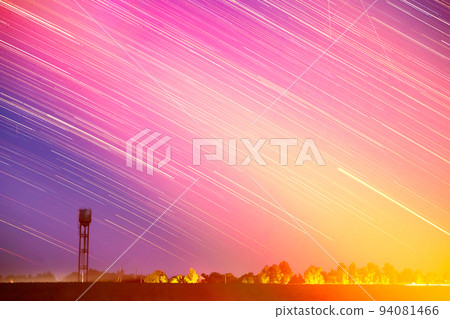 Amazing Unusual Stars Effects In Sky. Stars Lines Move Above Rural Landscape Water Tower In Background. Star Trails On Night Sky. Exposure. Soft Colors. Abstract Bright Yellow, Red, Pink Colors 94081466