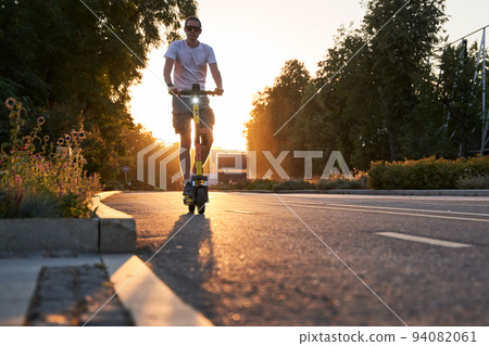 Man is riding on electric scooter or e-scooter on the street on the sunset. Man is riding on electric scooter or e-scooter on the street on the sunset. 94082061