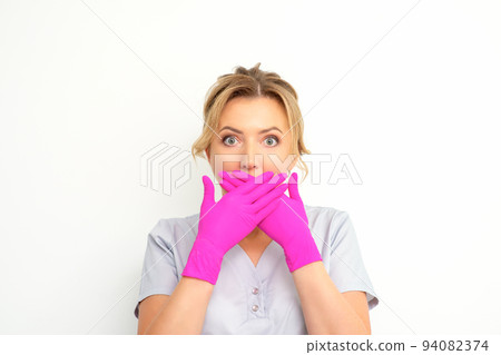 Portrait of a young female caucasian doctor or nurse is shocked covering her mouth with her pink gloved hands against a white background 94082374