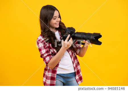 Positive and smiling emotions of teenager girl with dslr professional photo camera with big photo lens. Child photographer isoalted on yellow background. Photo school. 94085146