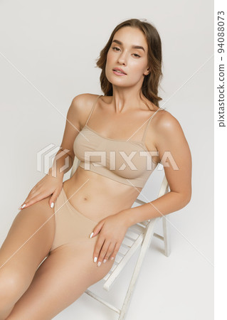 Young adorable smiling woman in lingerie sitting on floor isolated over gray background. Wellness, wellbeing, fitness and aesthetic cosmetology concept. Young adorable smiling woman in lingerie sitting on floor isolated over gray background. Wellness, wellbeing, fitness and aesthetic cosmetology concept. 94088073